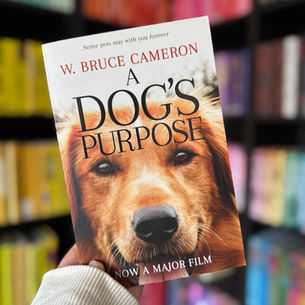 Hand holding "A Dog's Purpose" book by W. Bruce Cameron in a colorful bookstore. The cover shows a dog's face. Text: Now a Major Film.