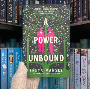 A gloved hand holds a book titled "A Power Unbound" by Freya Marske. The cover features green and pink designs. Bookshelves are in the background.