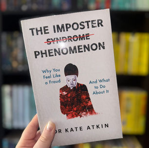 Close-up of a hand holding the book "The Imposter Phenomenon" by Dr. Kate Atkin, in front of a blurred bookshelf. The cover features a masked figure.