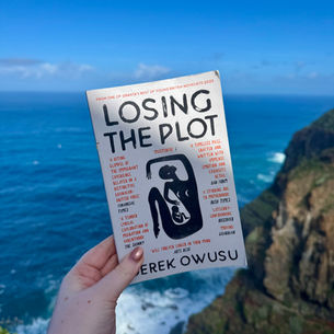 Hand holding the book "Losing the Plot" by Derek Owusu against a backdrop of ocean cliffs and blue sky.