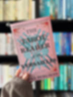 Hand holds "The Tarot Reader of Versailles" book against a bookshelf. Pink cover features intricate blue designs. Shelves filled with colorful books.