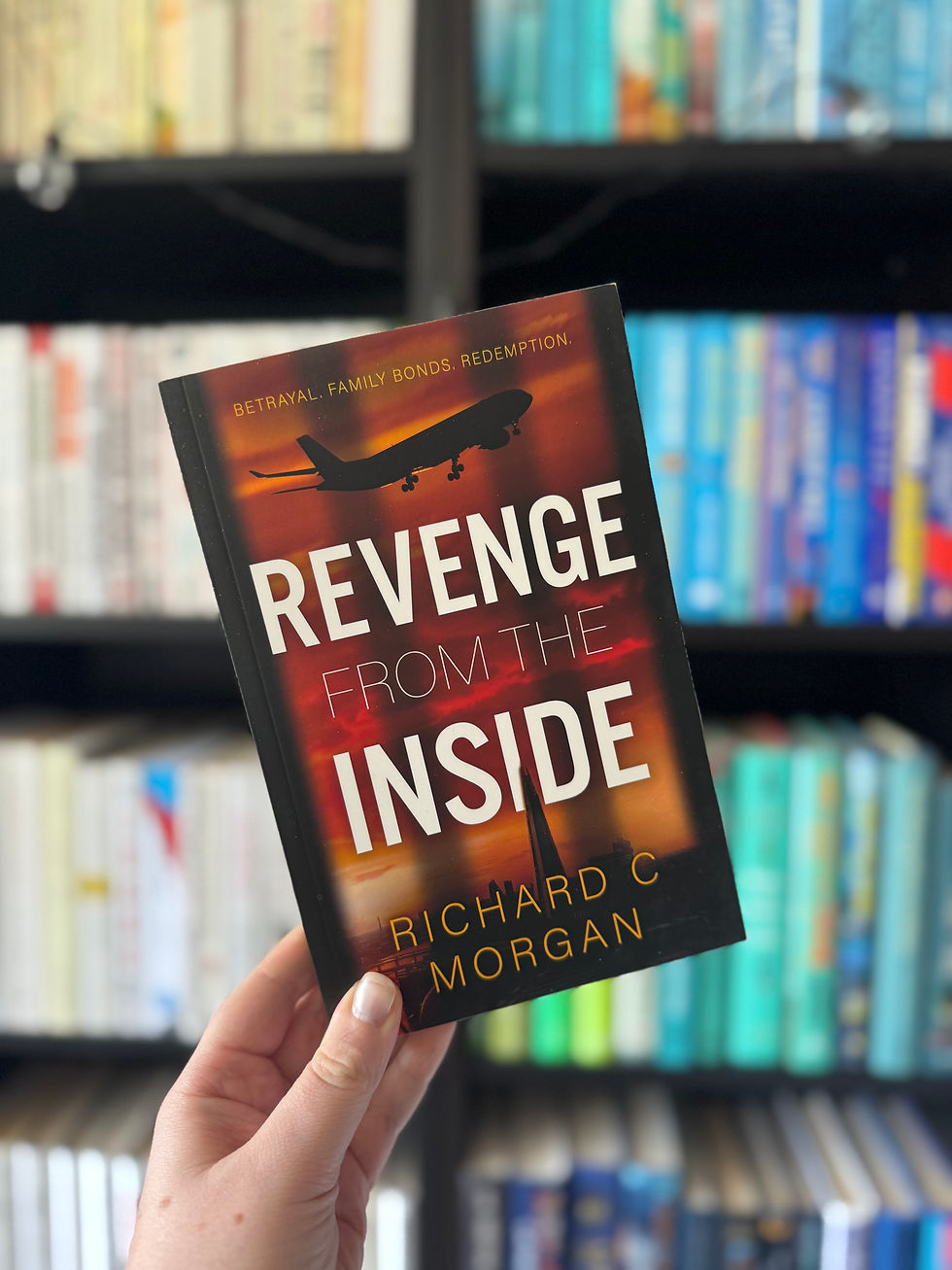 Hand holds book titled "Revenge from the Inside" by Richard C. Morgan. Background shows a colorful bookshelf. Airplane silhouette on cover.