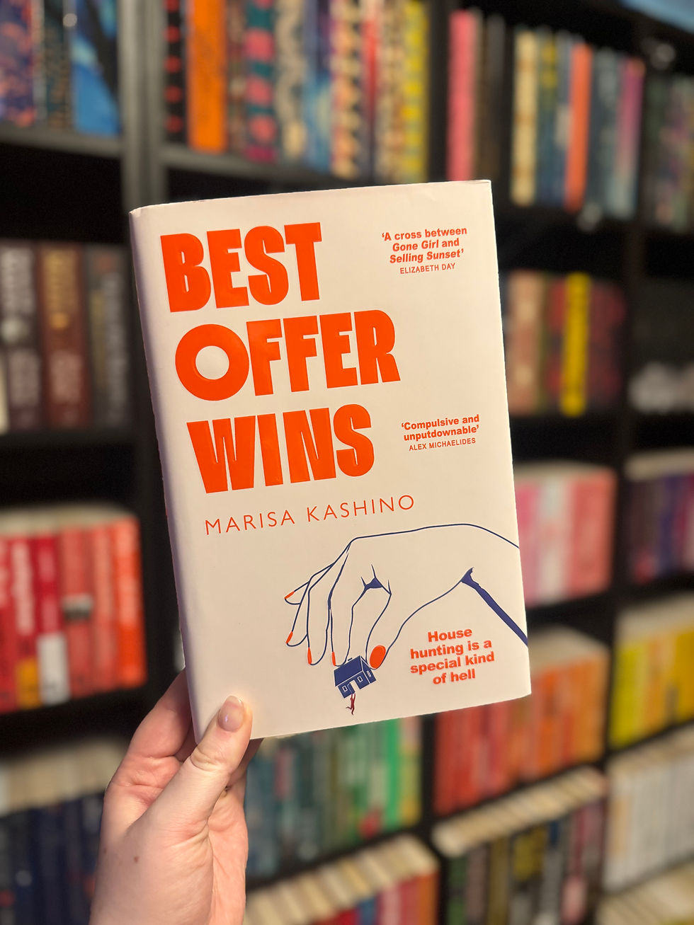 Hand holding a book titled "Best Offer Wins" by Marisa Kashino. Shelves of colorful books blur in the background, creating a cozy bookstore vibe.