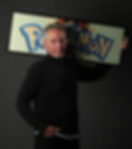 Man in a black sweater holds a framed Pokemon sign with colorful lettering against a dark background, exuding a confident demeanor.