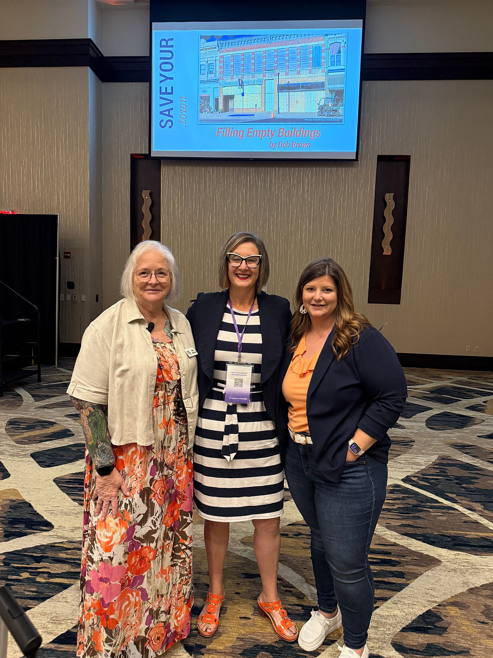 Three women stand together at the Kansas Entrepreneurship & Youth Summit beneath a presentation screen displaying “Filling Empty Buildings” by Deb Brown of SaveYour.Town.
