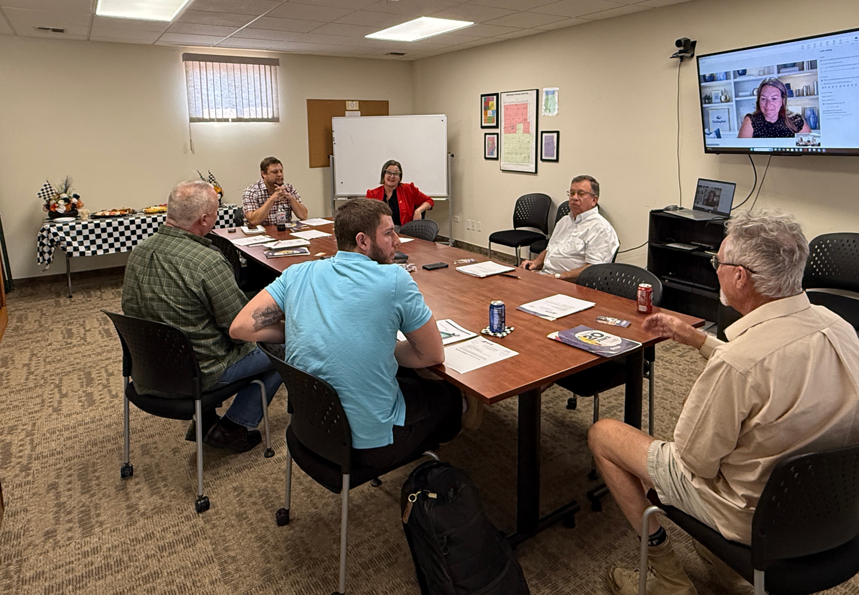 A group of local leaders and Network Kansas representatives sit around a conference table during Dickinson County’s eCommunity onboarding, with a remote participant appearing on a wall-mounted screen.