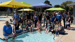 Do-Alogue participants gather around the shallow end of the Solomon Municipal Pool beneath colorful shade structures.