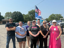 Seven Junction Point Post Acute employees stand together outdoors during the annual Car Show in Herington, Kansas, with vehicles and American and racing flags behind them.