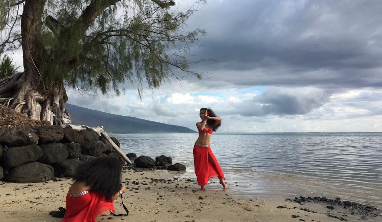 In this photo, a woman dances before a scenic seascape. We see the sand, ocean and a tree in the background, while the dancer makes an arm gesture crossing one hand over the other, palms flat.