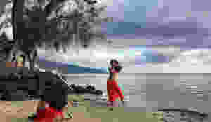 In this photo, a woman dances before a scenic seascape. We see the sand, ocean and a tree in the background, while the dancer makes an arm gesture crossing one hand over the other, palms flat.
