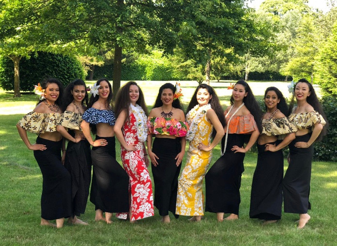 A group of dancers wearing black skirts and flower print tops stand in a line and smile at the camera.