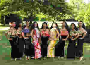 A group of dancers wearing black skirts and flower print tops stand in a line and smile at the camera.