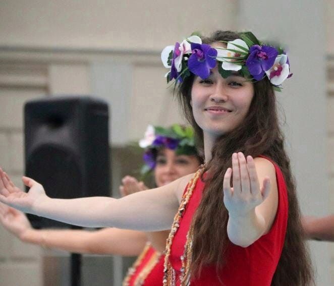 A girl with long brown hair stands side-on looking at the camera with her arms spread wide, hands facing upwards.