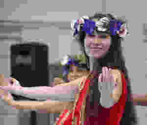 A girl with long brown hair stands side-on looking at the camera with her arms spread wide, hands facing upwards.