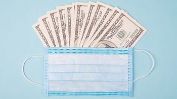 Fan of dollar bills behind a blue surgical mask on a light blue background, suggesting financial implications of healthcare.