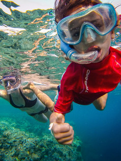 otrok v rdeči majici in z masko ter mama v črnih v ozadju  snorklata v prelepem morju