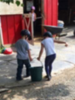 Image: Two children carrying a water bucket towards a barn stall.
