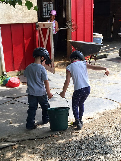 Image: Two children carrying a water bucket towards a barn stall.