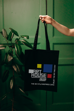 Hand holding black "ART HOUSE CAFE" tote bag, green wooden background.