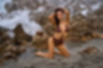 Woman in brown bikini poses on rocky beach, waves crashing behind. Dramatic and confident mood. Wet hair, gray skies, no visible text.