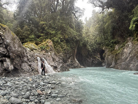 Hiker in black clothing standing alone by a glacial river deep in a green canyon