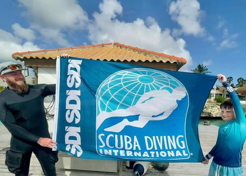 Man and child holding up a Scuba Diving International flag on wooden pier under bright blue sky