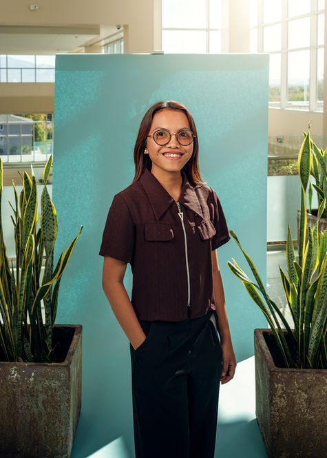 Professional portrait of a young woman with round glasses smiling in front of a teal backdrop with snake plants in a bright office atrium