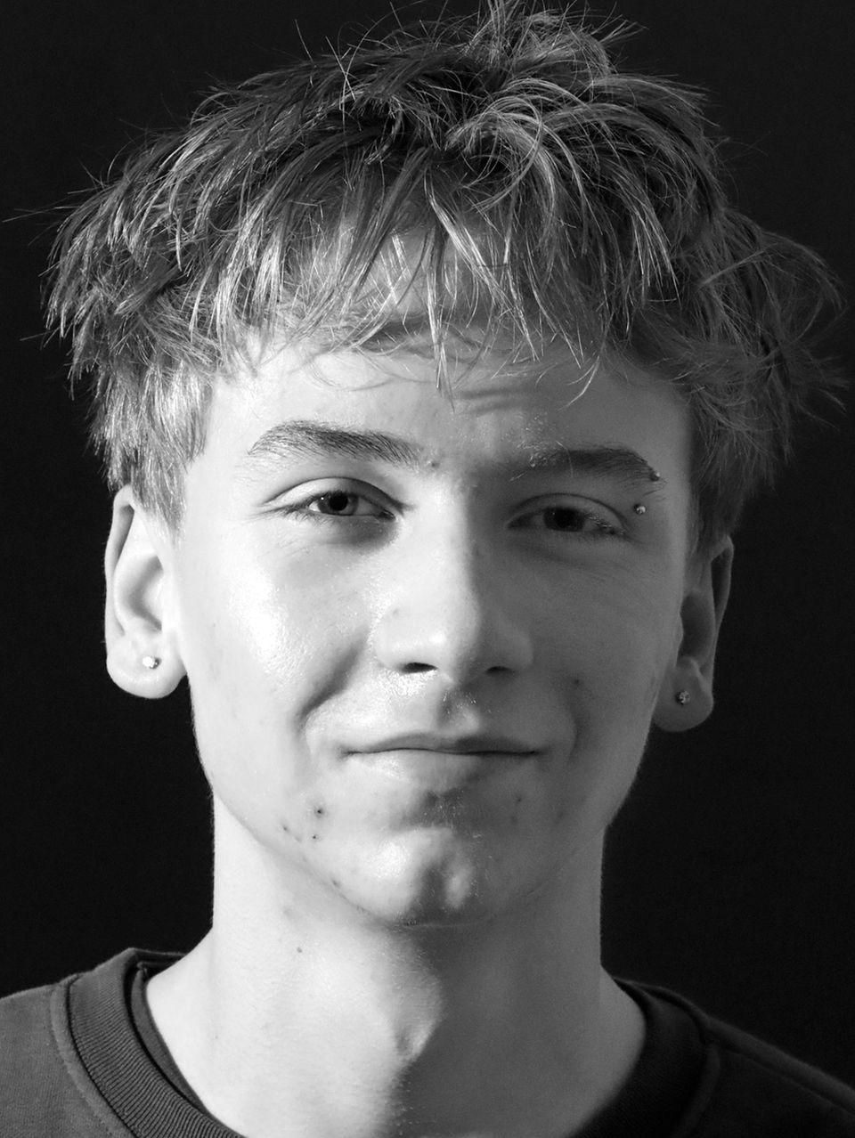 Junger Mann mit kurzen, zerzausten Haaren lächelt leicht in Schwarz-Weiß.