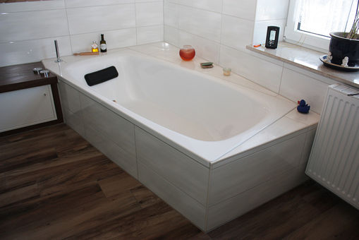Eckbadewanne mit weißer Verkleidung, Holzoptik-Boden, Ablage und Deko-Elementen am Fenster