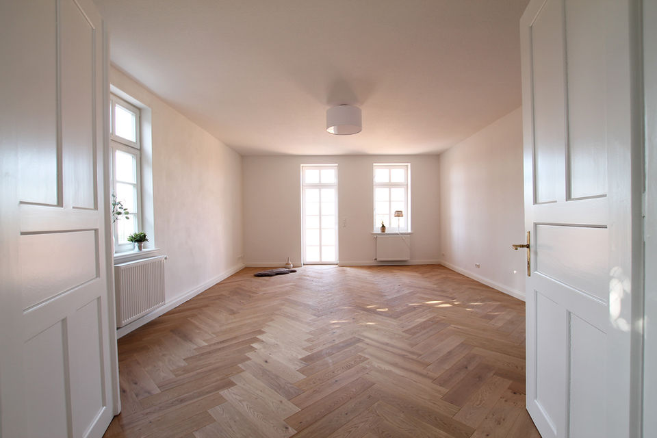 Leerer Raum mit weißen Wänden, Fischgrätparkett, großen Fenstern, offenen Türen.