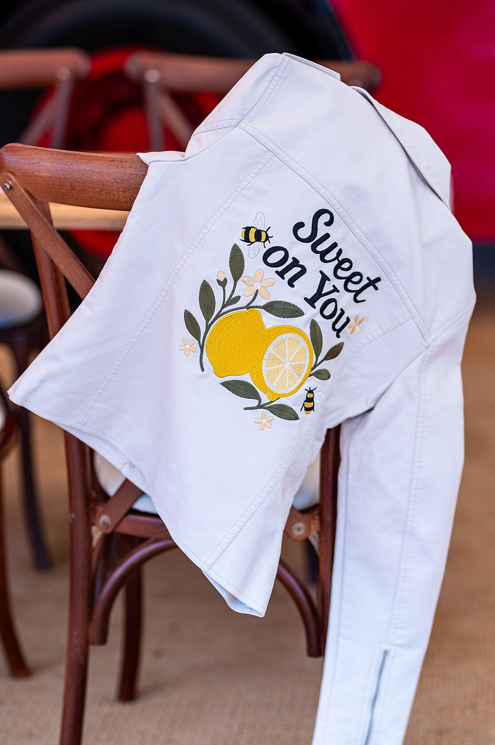 White jacket with "Sweet on You" and lemon graphic on chair. Red bus in background, bright paper decorations, warm atmosphere.