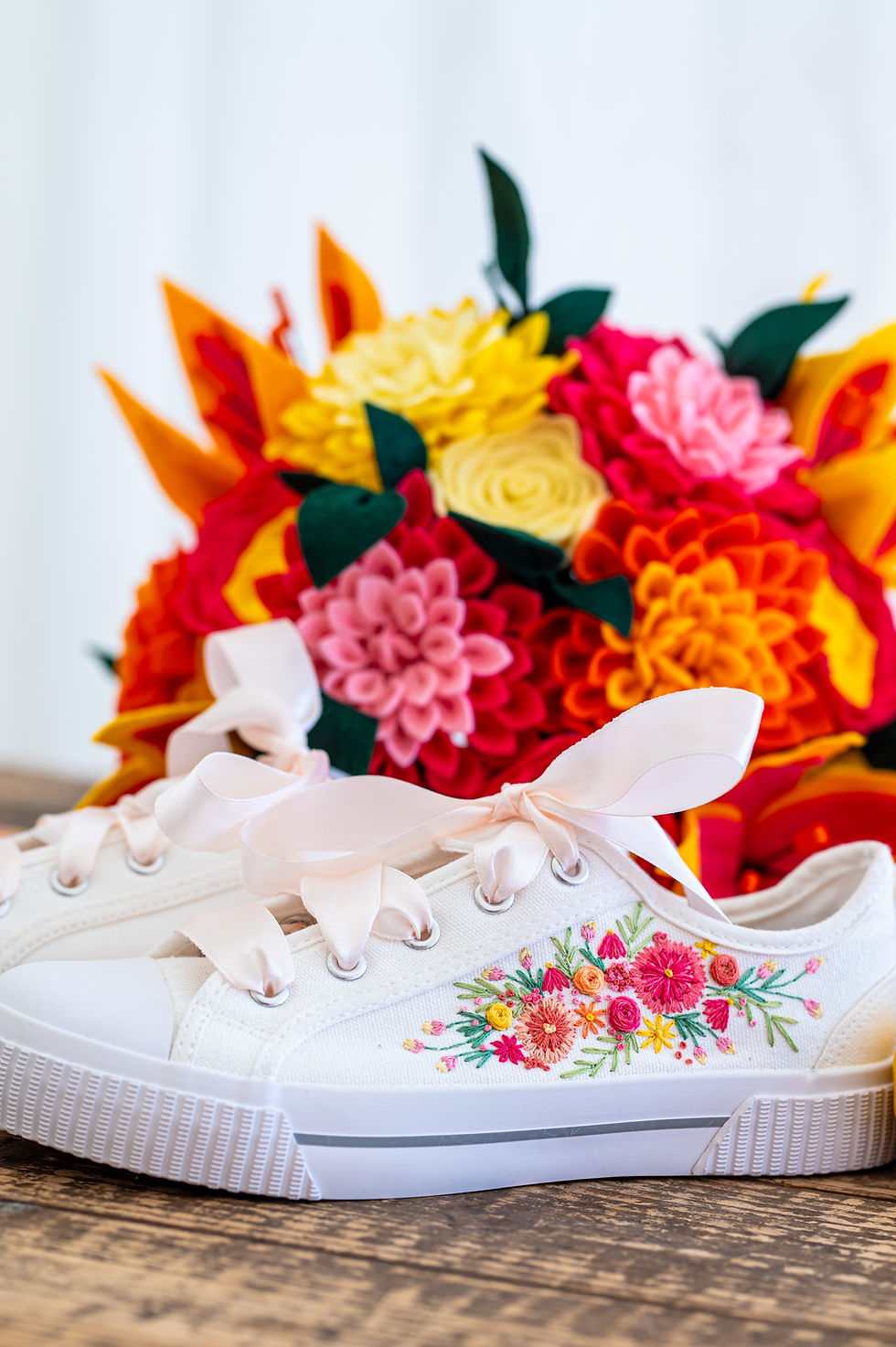 White sneaker with floral embroidery, pink ribbon laces on a wooden surface. Vibrant felt flower bouquet with red, yellow, and pink blooms.