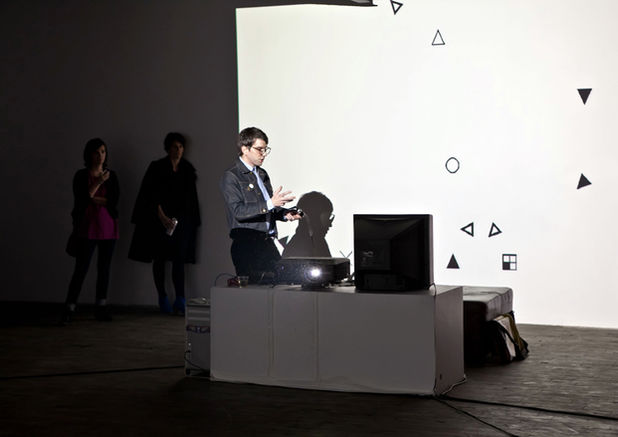 A group of people standing and sitting in front of a digital art installation with abstract shapes projected onto a wall. A person is working at a computer, interacting with the installation in a darkened gallery space.
