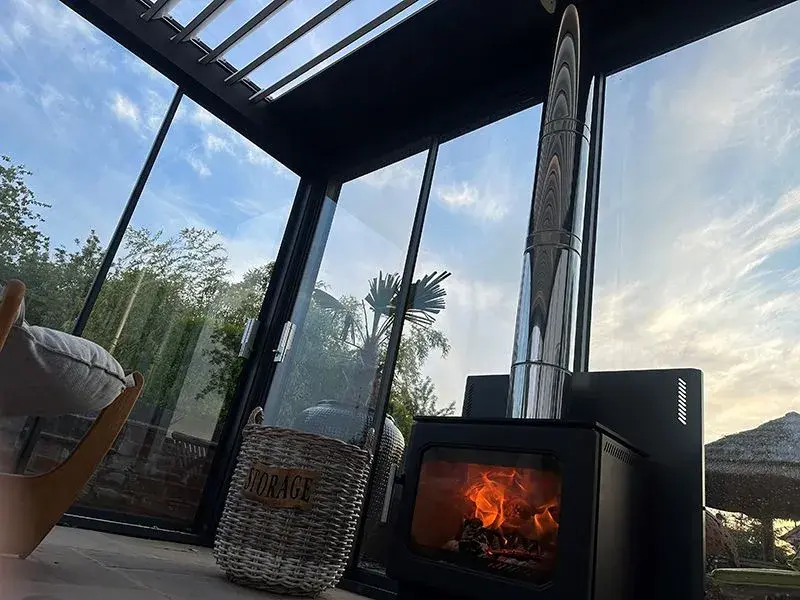 Close-up view of log burner outdoor heater glowing warmly inside the garden room made of bioclimatic pergola enclosed with sliding glass door.