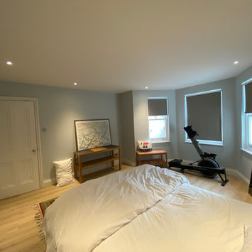 Modern bedroom with double bed, white bedding, wooden headboard, colorful rug, rowing machine, wooden chair, bedside table, desk, bay window with blinds, and wooden floor.
