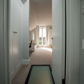 Bright attic hallway with white paneling, glass floor panel, and large windows.
