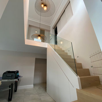Modern staircase with light wooden steps, glass railing, pendant light, white paneling, tiled floor, under-stair storage, and entryway table
