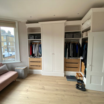 Modern dressing room with white built-in wardrobes, shelves, drawers, pink sofa bed, pillows, large window with curtains, wooden floor, and pile of shoes
