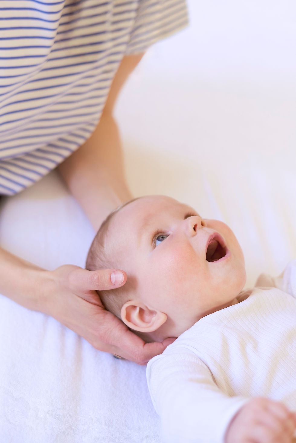 Baby in weißem Body auf dem Rücken auf Behandlungsliege, Hände der Osteopahin von h3 am Kopf des Babys, Handgriff aus der craniosakralen Osteopathie als Behandlugsansatz für Babys