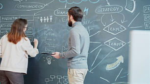 Two people collaborate at a chalkboard filled with strategic diagrams and names. The setting is a creative, brainstorming session.