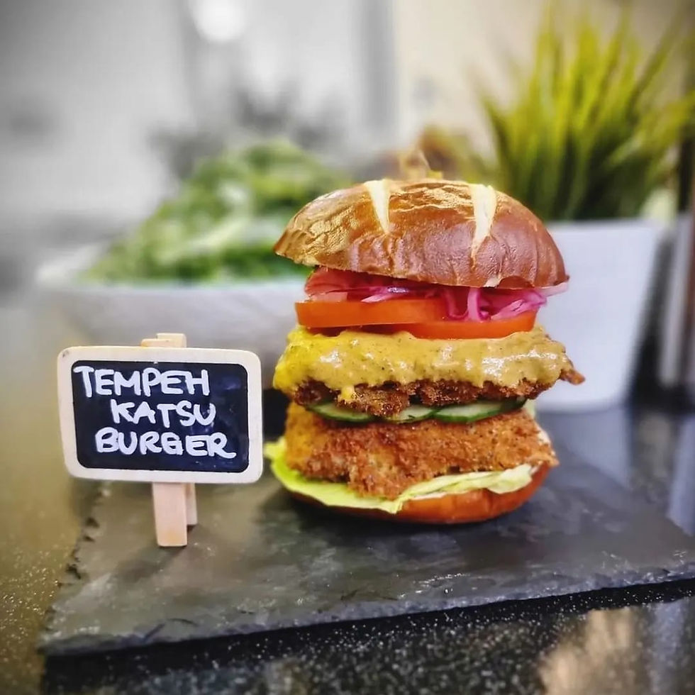 A stacked tempeh katsu burger in a toasted brioche bun with crispy breaded tempeh patties, tomato, pickled red onion and cucumber, beside a chalkboard sign reading Tempeh Katsu Burger.