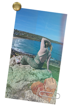 A photo of Carolyn, a white woman with blonde hair, sitting on a rock overlooking St Ive's harbor in Cornwall England. She is wearing a green lace gown she crocheted. The gown makes her look like a stunning goddess who is not to be trifled with. She is very proud. This photo is further edited to look like it is pinned to the corkboard background of this page. There is a brass tack in the upper left corner and a graphic of an origami heart "paperclipped" to the bottom of the photo.