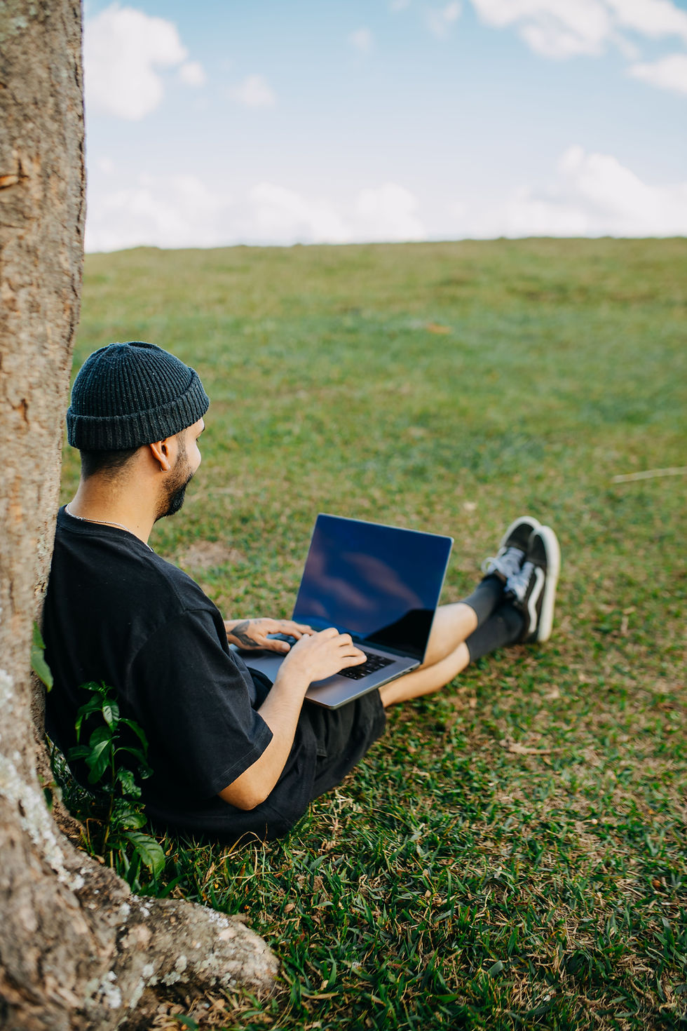 Hombre con gorro negro usa una laptop sentado en el césped junto a un árbol, bajo un cielo despejado y tranquilo.