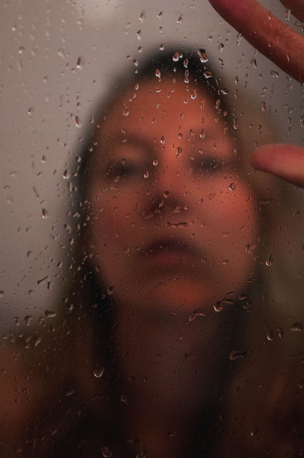 Vage vrouw achter met regendruppels beslagen glas, met vingers tegen het raam; sombere, introspectieve sfeer.