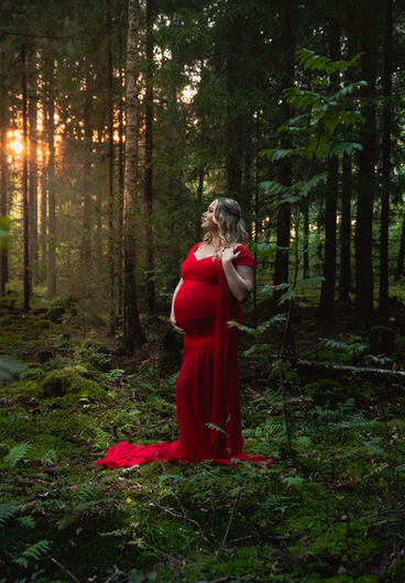 Gravid kvinna i lång röd klänning står i profil i en mossig skog med varmt kvällsljus mellan träden.