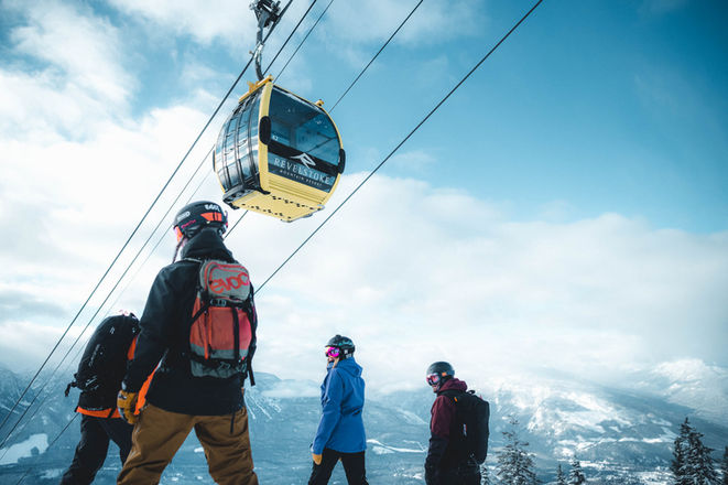 Skiers beneath a gondola with snowy mountains behind them