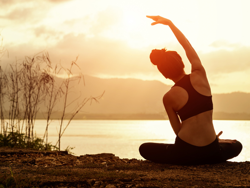 Woman practicing seated yoga pose at sunrise near a lake, stretching her arm overhead with mountains in the background — promoting mindfulness and flexibility through outdoor yoga.