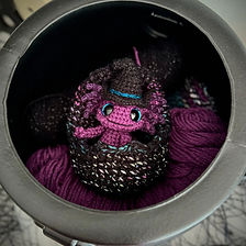 A small, purple axolotl wearing a witch's hat nestled inside a crocheted egg and yarn within a cauldron. 