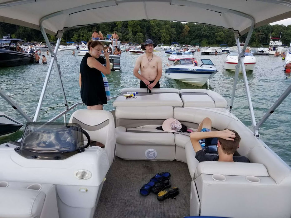 Group of people lounging on a pontoon boat at a crowded lake with many boats and people in the background enjoying a sunny day on the water.