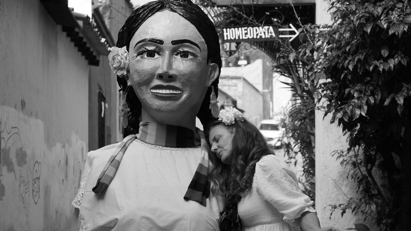 Black-and-white photo performance from “A Transformation Machine” by Canadian-Argentinian female artist Jazmín Sasky, featuring expressive movement and interaction with a giant doll, exploring personal growth, self-discovery, and inner child connection.